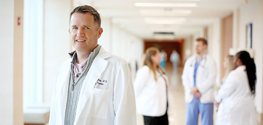 Headshot of Dr. McKay in a hospital hallway, with a group of Transitional Year residents chatting in the background.