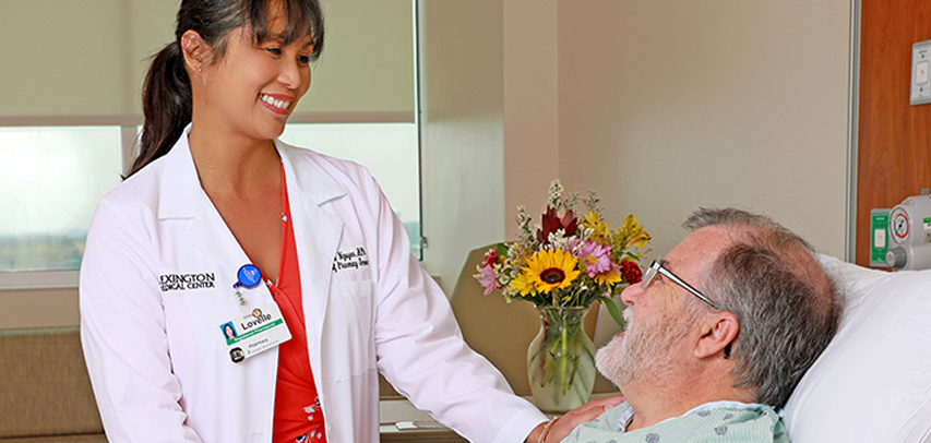A pharmacist at a patient’s bedside, with flowers on the side table in the background.