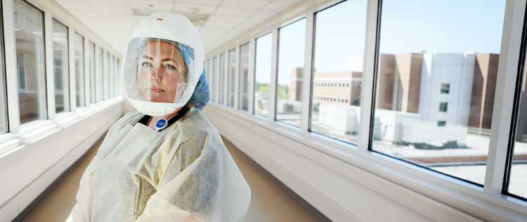An Acute/Critical Care nurse wears full scrubs and face shield as she stands patiently in a hallway lined with windows.