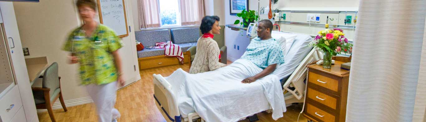 A Lexington Medical Center patient in a private room with a visitor, flowers on the bedside table, and nurse exiting the room in a blur of motion