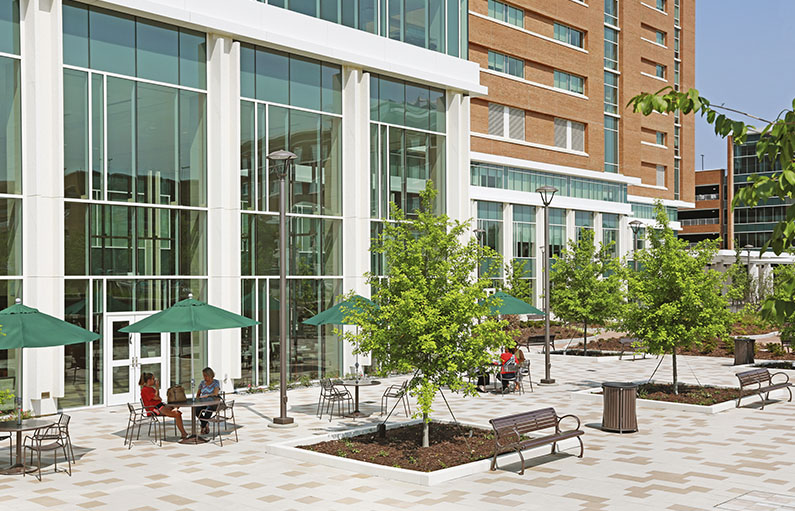 The courtyard outside the new LMC tower, landscaped with brick and trees, full of patio tables and chairs under green umbrellas.