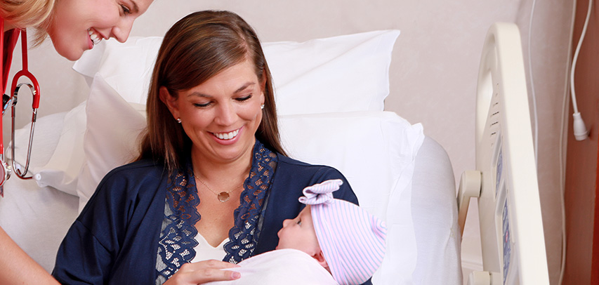 A woman in a hospital bed smiling down at her baby who is smiling back wide-eyed and mouth open, swaddled and wearing a pink cap with a front bow. A health care provider wearing a stethoscope is bending over the side of the bed smiling at both of them.