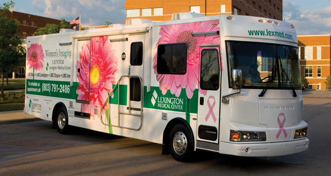 A large white bus with a green stripe down the side with white text that reads Lexington Medical Center Women’s Imaging Center LMC Irmo LMC Chapin LMC Lexington LMC. To schedule an appointment call 803-791-2486. Large pink flowers and pink breast cancer awareness ribbons decorate the rest of the truck. The front of the bus has green text saying www.lexmed.com.