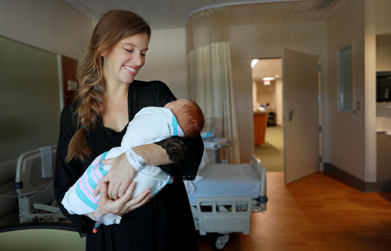 A woman standing in a delivery room holding and smiling down at her newborn baby swaddled in a blanket with a view of the open delivery floor hallway behind.