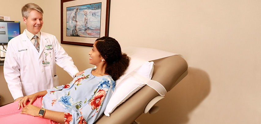An LMC doctor smiling with a female patient who is on a medical recliner in a warm examination room with art on the wall. 