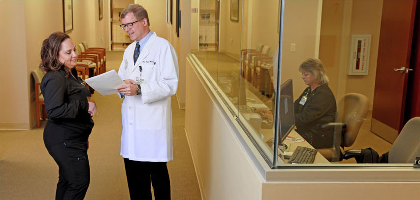 An LMC doctor and nurse standing in a warm doctor’s office hallway lined with chairs and art, looking over paperwork together.